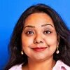 Person looks directly at the camera, long dark hair, earrings, blue background.