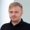 Dr. Hendrik Simon Mann mit kurzem blondem Haar und Bart schaut in die Kamera, steht vor heller, unscharfer Wand.
