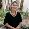 Ana Cláudia Duarte Cardoso Person with short wavy gray hair, glasses, and green necklace sits outdoors smiling, hands resting on patterned trousers.