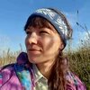 Person looks to the left, wears a patterned headband, and stands outdoors with dry vegetation and blue sky in the background.