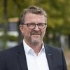hbs Bildbeschreiber sagte:  Person mit Brille steht draußen vor unscharfem Hintergrund mit Bäumen und Wegen.