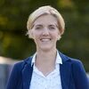 Person mit kurzem, blondem Haar steht draußen und blickt lächelnd in die Kamera. Unscharfer Hintergrund mit Bäumen.