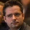 Ein Mann mit Brille schaut ernst zur Seite, im Hintergrund sind verschwommene Personen.