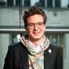 Person mit Brille lächelt in Innenraum vor Fensterfront und blickt in die Kamera.