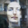Nahaufnahme eines Gesichts mit blasser Haut und dunklem Haar. Person schaut ernst durch eine leicht reflektierende Scheibe.