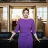 Person mit blonden Haaren und violettem Kleid steht vor einem Tisch, stützt sich mit beiden Händen ab, schaut in die Kamera; im Hintergrund geöffnete Tür und Besprechungsraum.