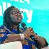 Fatou Warkha Sambe, wearing glasses and a patterned blue blouse, sits on a stage, holding a microphone and speaking in front of a turquoise screen.