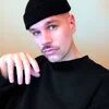 Person mit schwarzer Mütze und Schnurrbart blickt in Kamera, Hand im Vordergrund, neutraler Hintergrund.