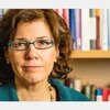 Porträt einer Frau mit Brille, frontal, ernster Blick. Im Hintergrund unscharfe Bücherregale.