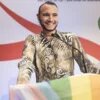 A smiling person with short dark hair and a beard stands at a lectern with a microphone, above which hangs a rainbow flag.