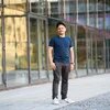 Eine Person mit kurzem Bart und Schnurrbart steht aufrecht vor einer Glasfassade, trägt Kappe, T-Shirt, Hose und Sneaker, blickt ernst zur Kamera.