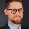 Person mit Brille und Bart blickt zur Kamera, trägt Hemd, Krawatte und Sakko, neutraler Hintergrund