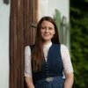 Frau in Tracht lehnt an Holzwand und lächelt in die Kamera, im Hintergrund Grünpflanzen.