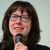 Frau mit Brille spricht lächelnd in ein Mikrofon vor hellem Hintergrund.