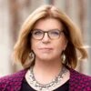 Frau mit Brille und Schmuck blickt frontal in die Kamera, Hintergrund unscharf.