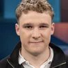 Junger Mann mit lockigen Haaren vor blauem Hintergrund, leicht lächelnd.