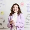 Lächelnde Person vor Whiteboard mit Notizen und Haftzetteln, gestikulierend im Gespräch.