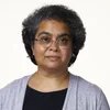 Person mit Brille blickt frontal in die Kamera, kurze lockige Haare, neutraler Gesichtsausdruck, heller Hintergrund.