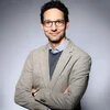 Lächelnde Person mit Brille blickt in die Kamera, Arme verschränkt, vor neutralem Hintergrund.