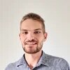 Lächelnde Person mit kurzem Haar und Bart blickt in die Kamera, vor heller Wand.