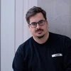 Portrait Kristian Brakel, Person mit kurzen Haaren und Brille in schwarzem Pullover vor hellgrauer Wand, schaut direkt in den Kamera.