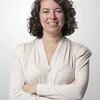 Eine lächelnde Frau mit schulterlangen, dunklen Locken steht mit verschränkten Armen vor einem hellen Hintergrund.