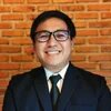 Photo: A person with glasses and a tie in a dark suit, smiling in front of a brick wall.