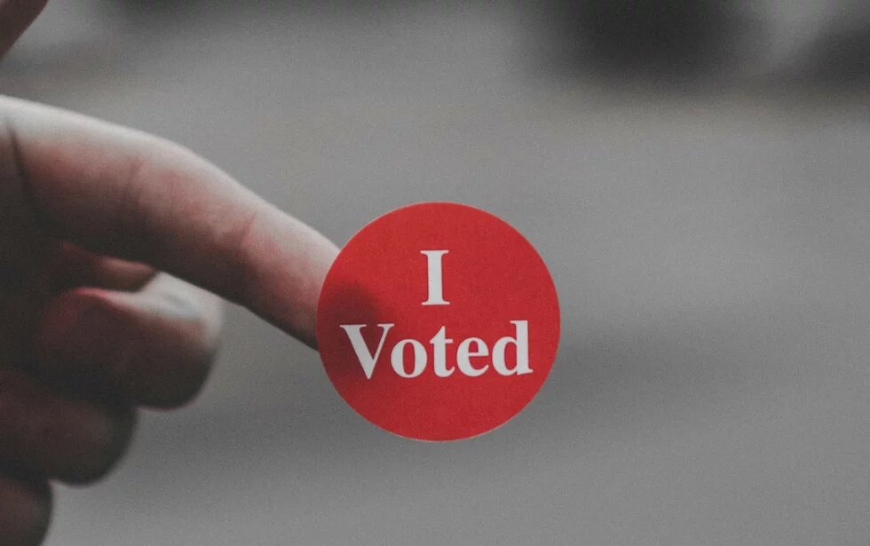 One hand is holding a sticker with "I Voted" written on it against a blurred background.