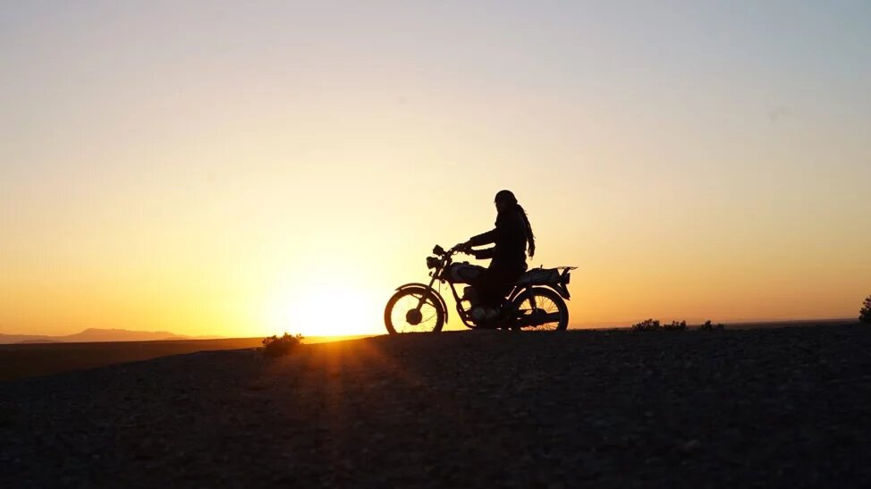 Zu sehen ist die Silhouette eines Menschen auf einem alten Motorrad, dahinter die untergehende Sonne