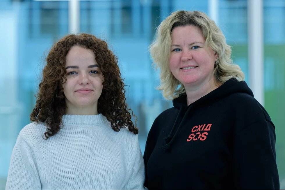 Yuliia Matviichuk (braune Locken, weißes Oberteil) und Oksana Kuiantseva (schwarzer Kapuzenpulli und blond gefärbte Haare) stehen nebeneinander und lächeln in die Kamera
