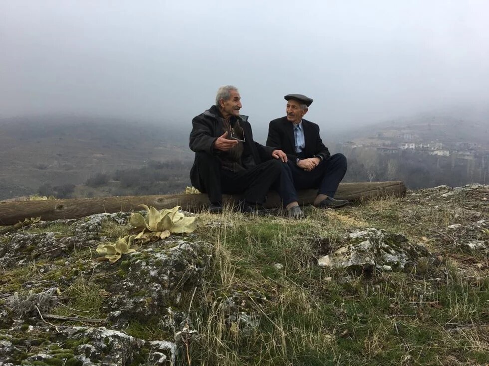 Asadur: Die Suche nach verlorener Identität zwei alte Männer sitzen auf einem Holzstamm auf einem Berg und unterhalten sich angeregt.
