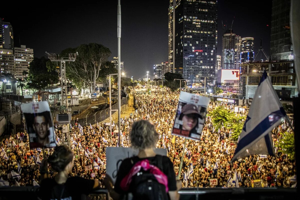 Demonstration in Tel Aviv, 22. Juni 2024. Foto: Nachtaufnahme in Tel Aviv, viele Demonstrant*innen mit Fahnen und Plakaten auf einer Straße, Wolkenkratzer im Hintergrund.