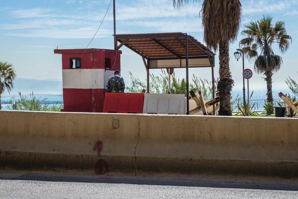 Militär-Checkpoint in Beirut Foto: Ein Kontrollpunkt am Meer, ein uniformierter Mensch sitzt in einem rot-weißen Wachhäuschen, Palmen im Hintergrund.
