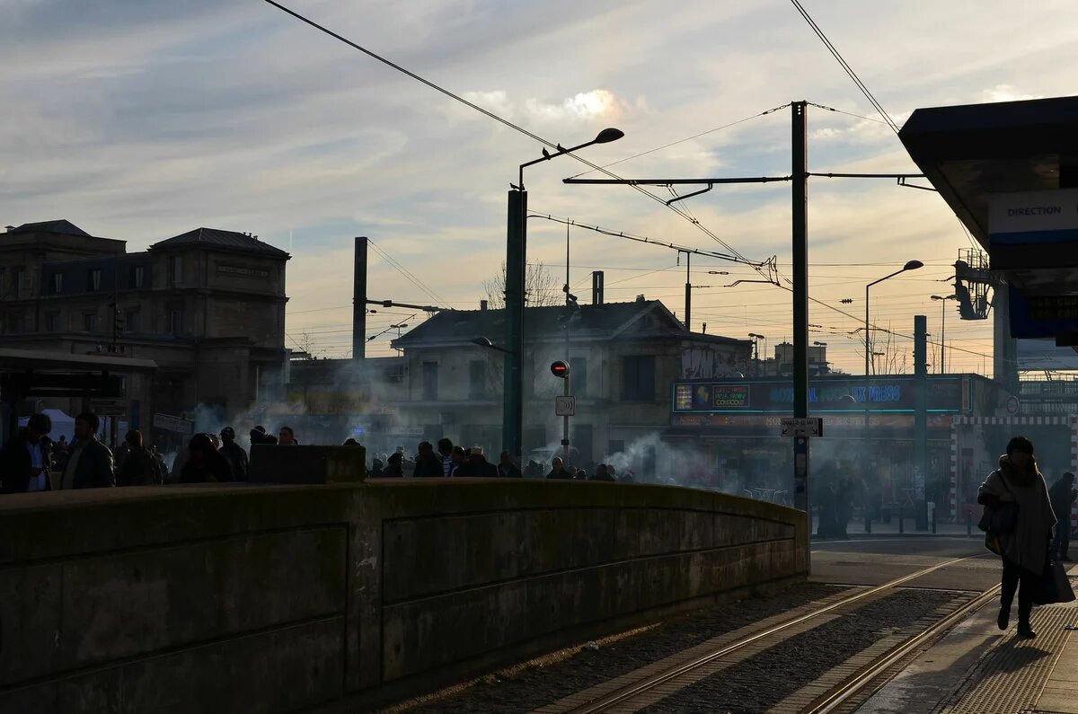 GPT Foto: Eine Menschenmenge steht auf einem Platz vor dem Bahnhof Saint-Denis in Paris, umgeben von Straßenbahnschienen und Oberleitungen. Rauch steigt auf.