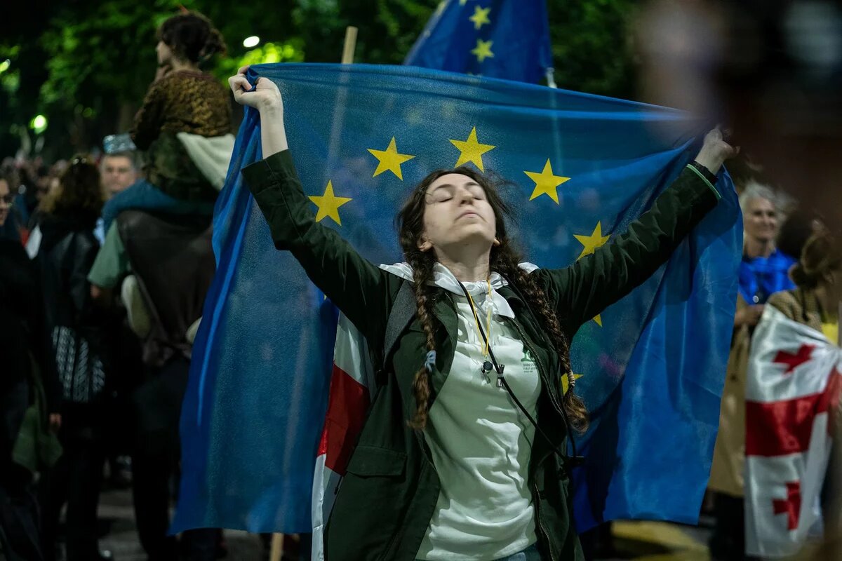 Tbilisi Protests 2024 Eine Person mit geschlossenen Augen hält eine EU-Flagge über den Schultern, umgeben von anderen Demonstrierenden, teils mit georgischen Flaggen.