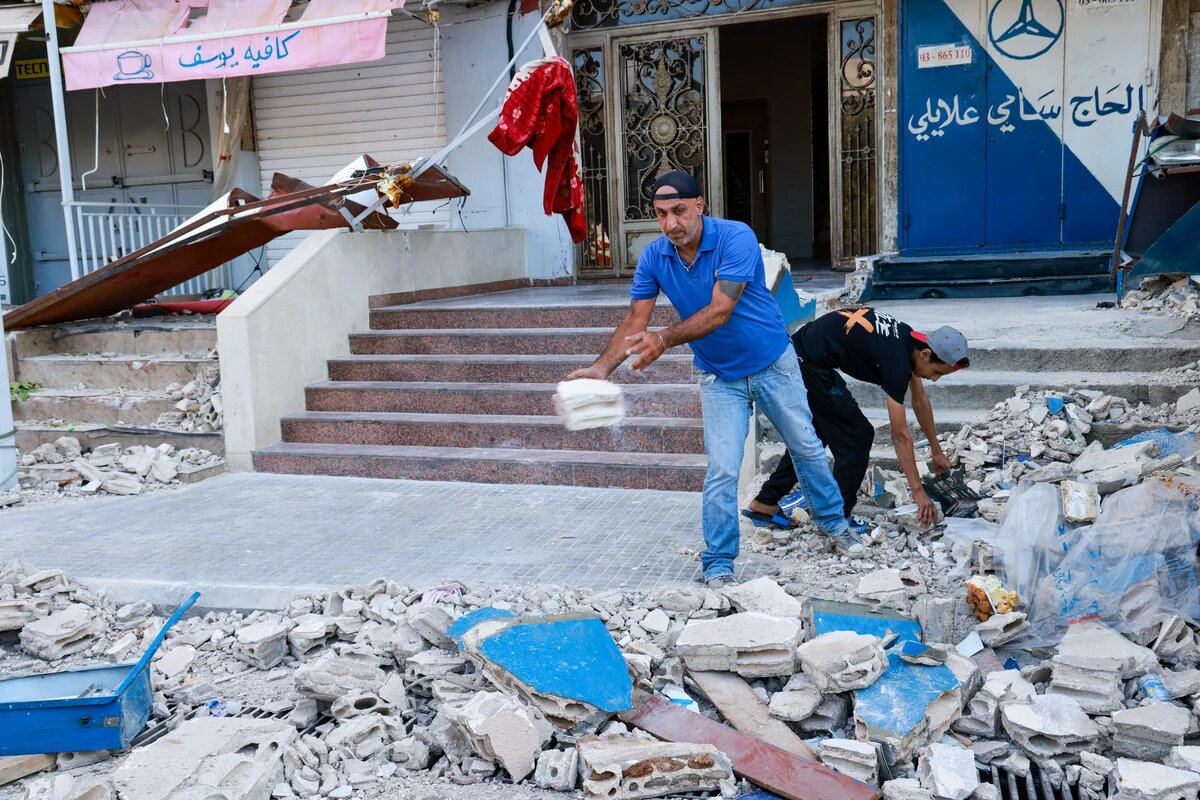 Aufräumarbeiten nach einem Drohnenangriff auf ein Gebäude im Cola-Viertel im Zentrum von Beirut am 30. September 2024 Foto: Zwei Männer räumen Trümmer von einer zerstörten Treppe vor einem Geschäft weg. Einer wirft Schutt, während der andere Trümmer aufhebt.