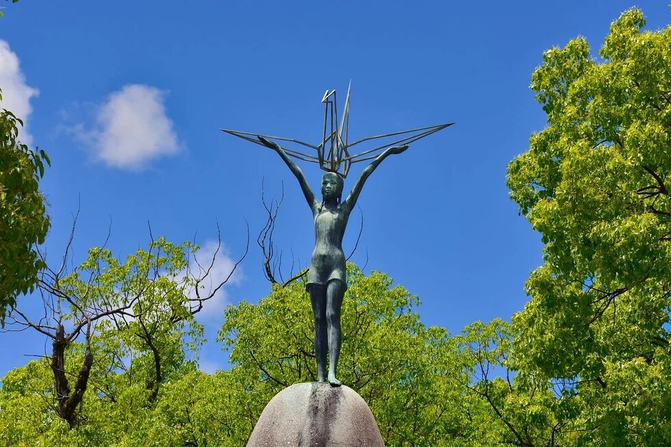 Children's Peace Monument in Hiroshima, Japan Photo: Statue of a child with outstretched arms holding a crane. Surrounded by trees, blue sky in the background.