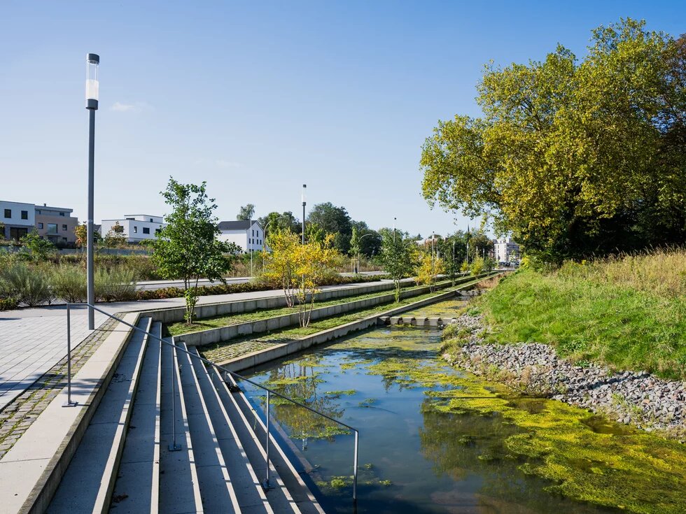 Stufenanlage führt zu einem bepflanzten Wasserbecken, gesäumt von Laternen und modernen Gebäuden im Hintergrund.