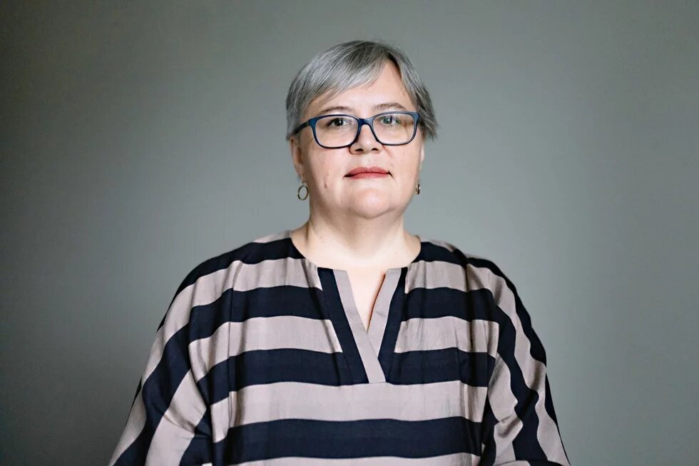 Irina Alkhovka GPT Icon Foto: Person mit grauem Haar und Brille vor neutralem Hintergrund, trägt ein gestreiftes Oberteil.