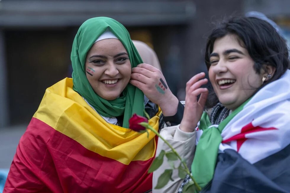 Syrische Frauen feiern in Duisburg das Ende des Assad-Regimes. Foto: Zwei lachende Personen mit bemalten Gesichtern und Fahnen um die Schultern, eine hält eine Rose in der Hand.