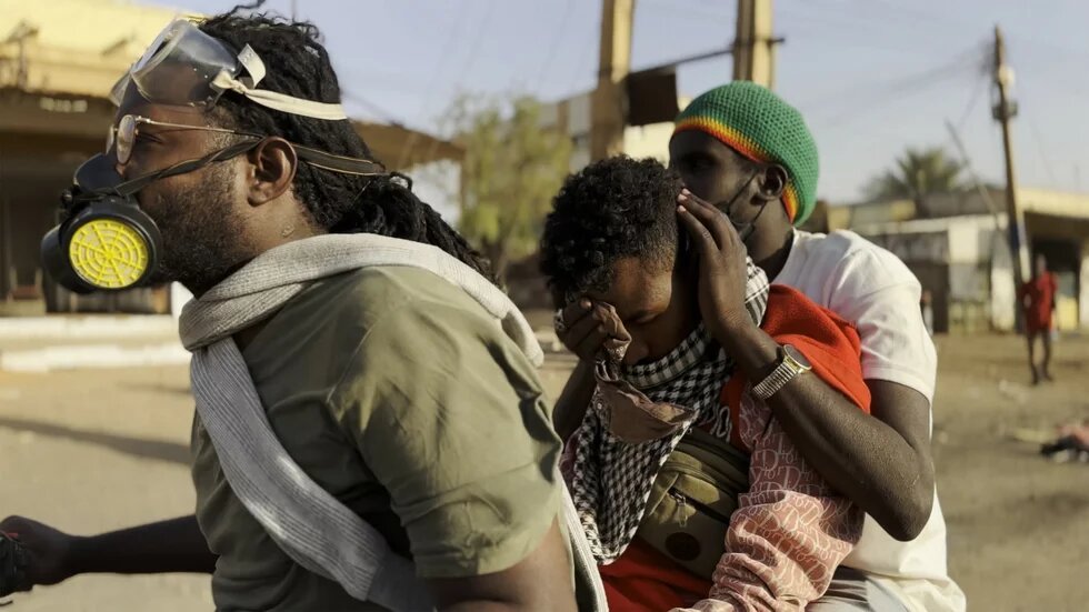 Jawad "Khartoum" Drei Personen auf einem Motorrad in einer staubigen Umgebung. Die vordere Person trägt eine Schutzmaske, die mittlere hält ein Kind, das das Gesicht bedeckt.