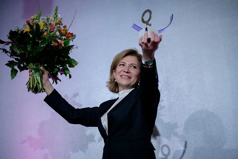 Joumana Seif, Anne-Klein-Frauen-Preis Joumana Seif hält den Anne-Klein-Frauenpreis in einer Hand und einen Blumenstrauß in der anderen. Sie lächelt vor einem neutralen Hintergrund.