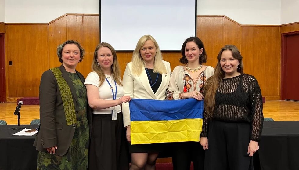 Foto: Fünf Personen stehen in einem Saal vor einer Leinwand und halten gemeinsam eine ukrainische Flagge.