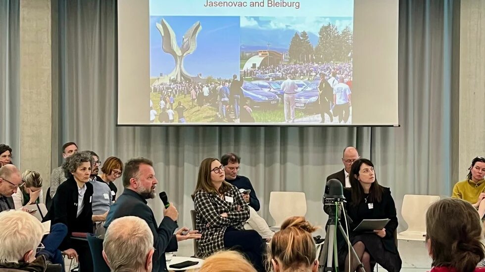 Mehrere Personen sitzen in einem Raum mit Stuhlkreis. Eine Person spricht ins Mikrofon. Im Hintergrund zeigt eine Leinwand Bilder mit dem Titel „Jasenovac and Bleiburg“.