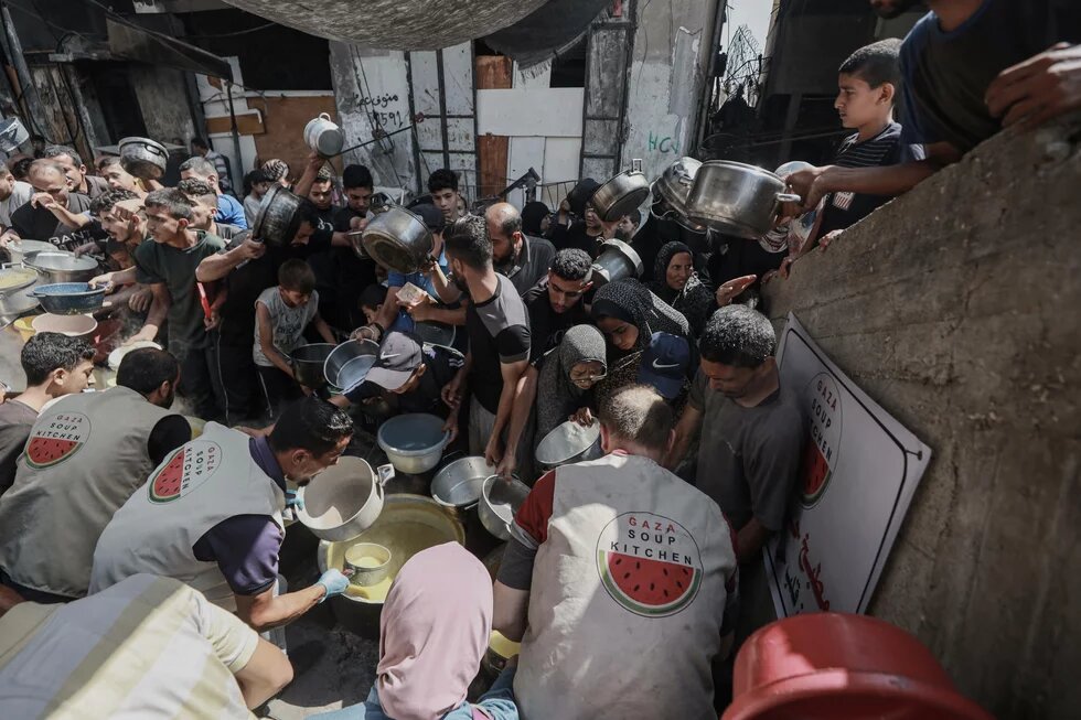 Gaza Soup Kitchen Viele Menschen mit Töpfen drängen sich vor großen Töpfen, aus denen Helfer*innen Suppe schöpfen (Gaza Soup Kitchen).