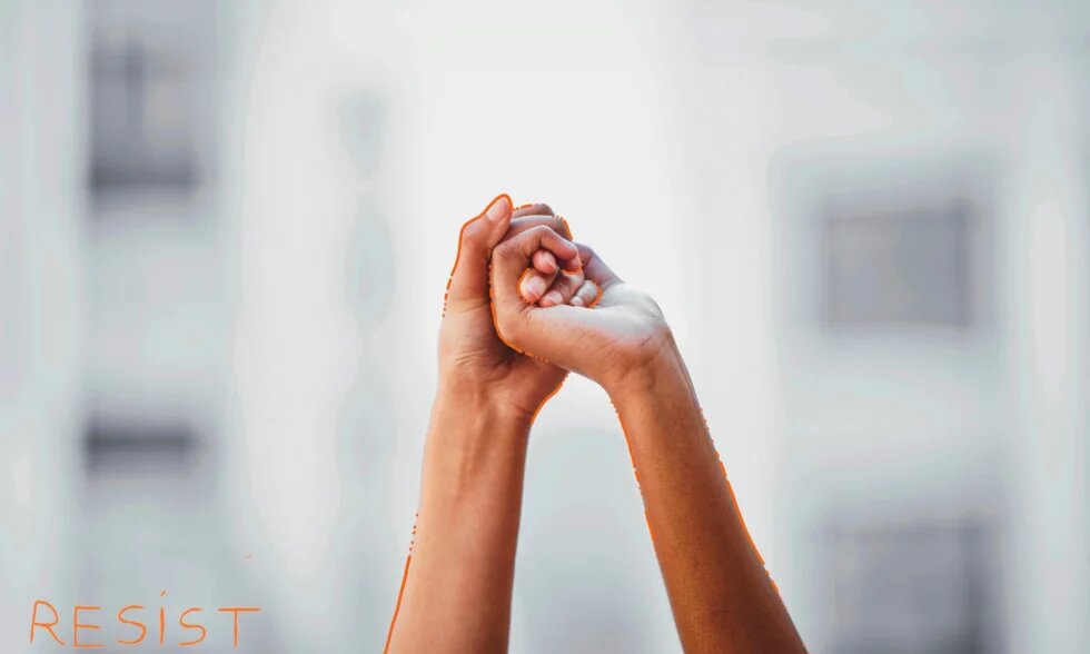 Two raised hands clasp each other, blurred buildings in the background; ‘RESIST’ is written at the bottom.
