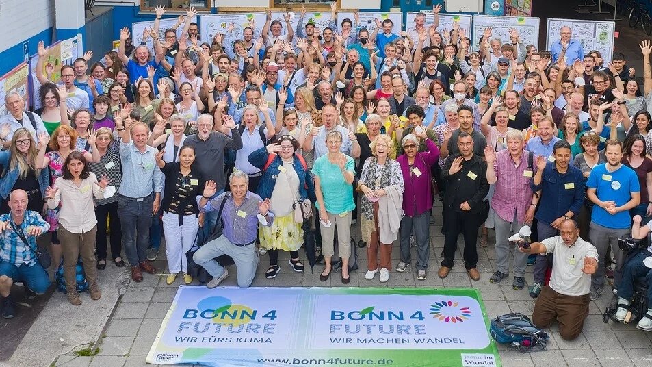 Zukunft beginnt vor Ort: Bonn4Future - Klimaforum Große Gruppe von Menschen winkt in die Kamera, stehend hinter zwei Bannern mit Aufschrift „Bonn4Future – Wir machen Wandel“.