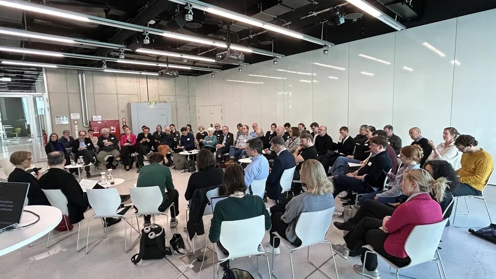 Frühjahrstagung 2025 der Grünen Akademie Über 50 Personen sitzen im Stuhlkreis in einem modernen Raum und hören einer Diskussion zu. Zwei Personen sprechen mit Mikrofon.