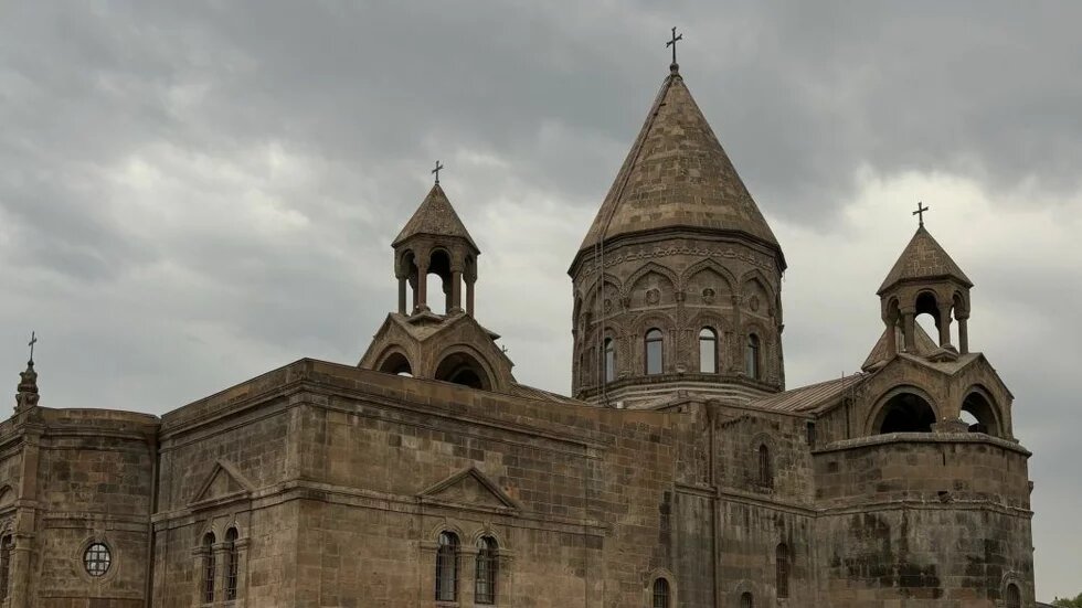 Steinerne Kirche mit drei spitzen Türmen und Kreuz, rechteckiger Baukörper, bewölkter Himmel im Hintergrund.