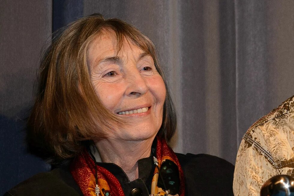 Ruth Marianne Wündrich-Brosien Eine ältere Person mit schulterlangem Haar lächelt, steht vor einem grauen Vorhang und trägt ein gemustertes Tuch.